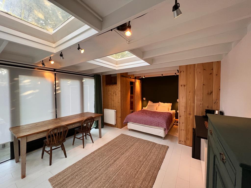 An open studio space features a spacious bed with warm bedding against a wooden accent wall. A dining area is present with a natural wood table and two chairs, while large skylights allow ample natural light to fill the room.