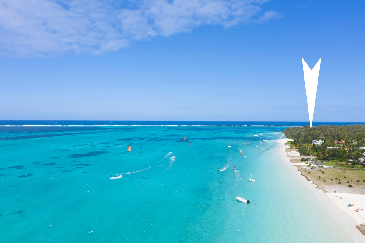 A panoramic view of a turquoise lagoon is presented, dotted with small boats and water sports activity. A sandy beach fringes the shoreline, bordered by lush greenery. The sky is clear with a few clouds, enhancing the sense of tranquility and open space.
