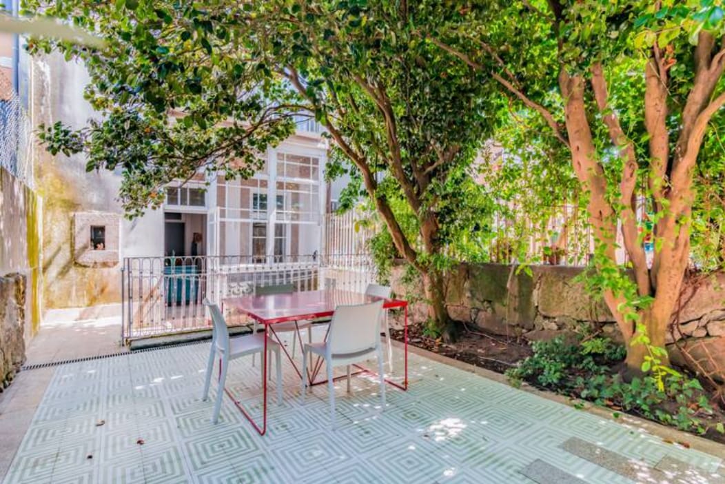 A common garden area features a spacious outdoor dining table with four chairs, surrounded by lush greenery. The area is adorned with mature trees, providing shade and a tranquil environment. A decorative tiled floor adds texture to the space, enhancing the garden's charm.