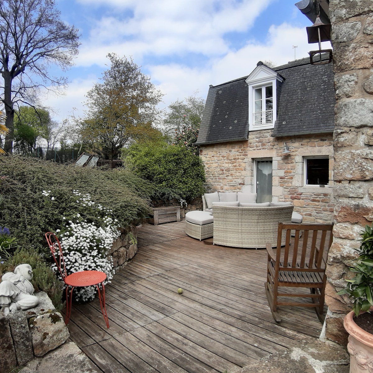 An inviting outdoor seating area is presented on a wooden deck, surrounded by lush greenery and flowering plants. Comfortable rattan furniture is arranged for relaxation, complemented by a small red chair. A stone wall creates a quaint backdrop against the charming architecture of the house.