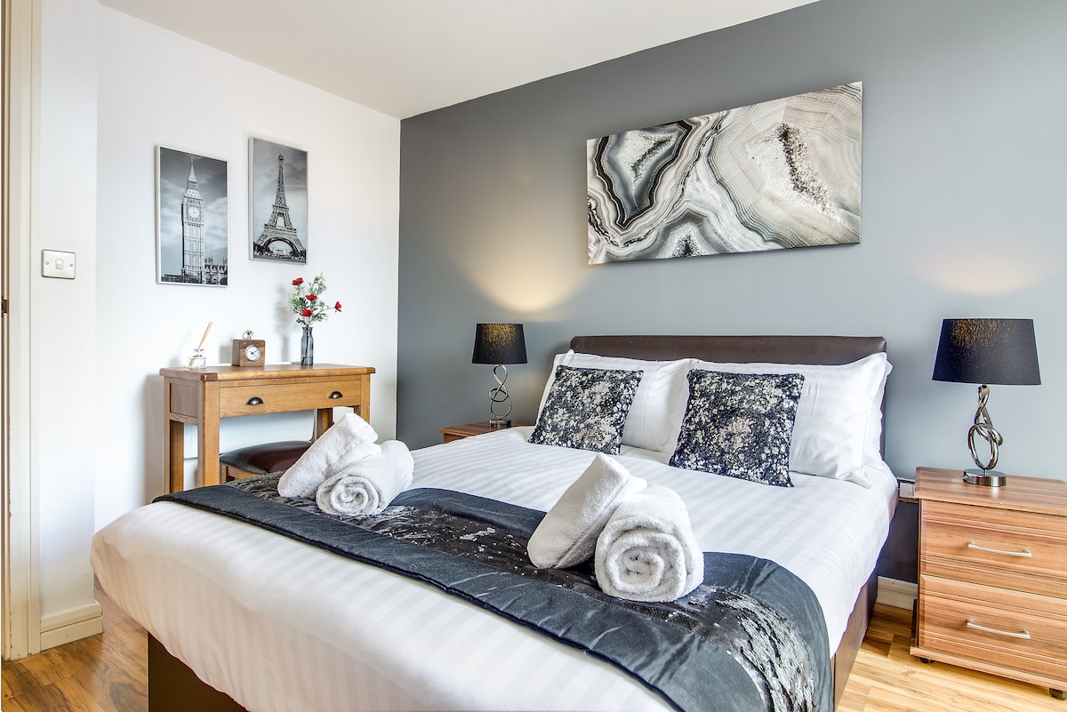 A well-appointed bedroom features a spacious bed adorned with neatly arranged towels. Subtle wall art and decorative lamps complement the room, while a wooden desk offers additional functionality. Natural light enters through a window, creating a tranquil setting.