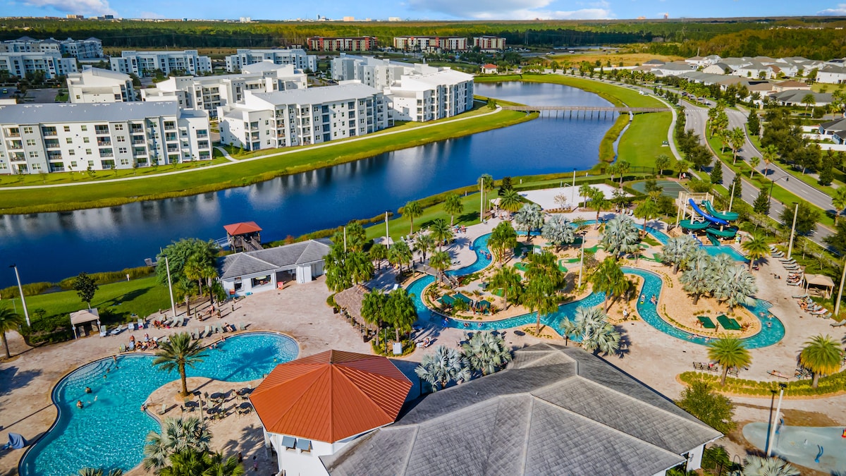 An aerial view showcases the expansive resort area, featuring a sprawling swimming pool surrounded by tropical landscaping. The scenic pond reflects the nearby apartments and lush greenery, while a waterslide and play areas are visible in the distance, offering various recreational options.