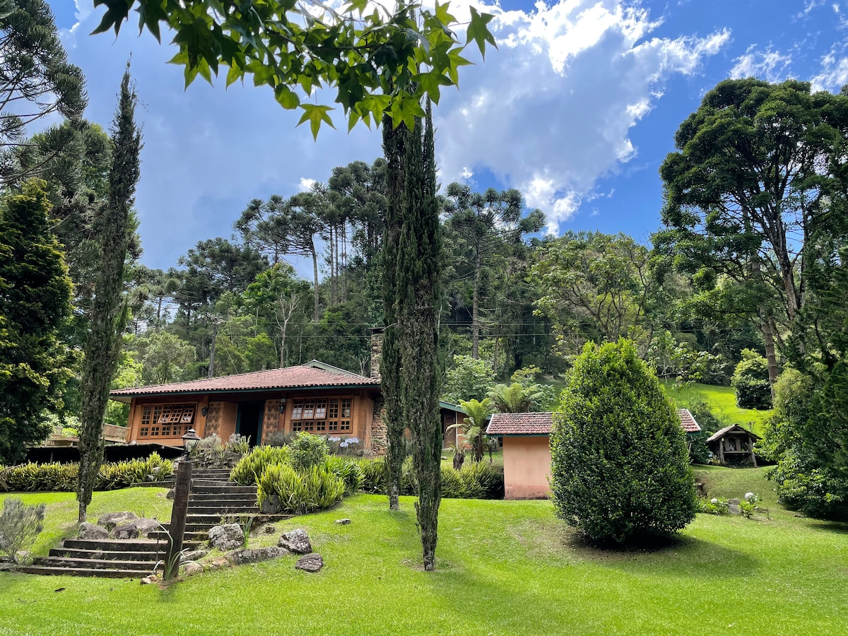 A charming wooden cabin is framed by lush greenery and tall trees. Steps lead to a well-manicured lawn, where vibrant shrubs and plants add to the natural surroundings. The sky is partly cloudy, creating a serene and inviting atmosphere.