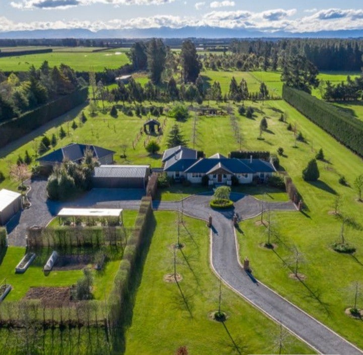 An aerial view showcases the expansive five-acre grounds of Silk Tree Cottage. Lush green lawns are interspersed with various trees and garden beds, while the modern cottage and surrounding buildings are framed by a winding driveway leading into the property.