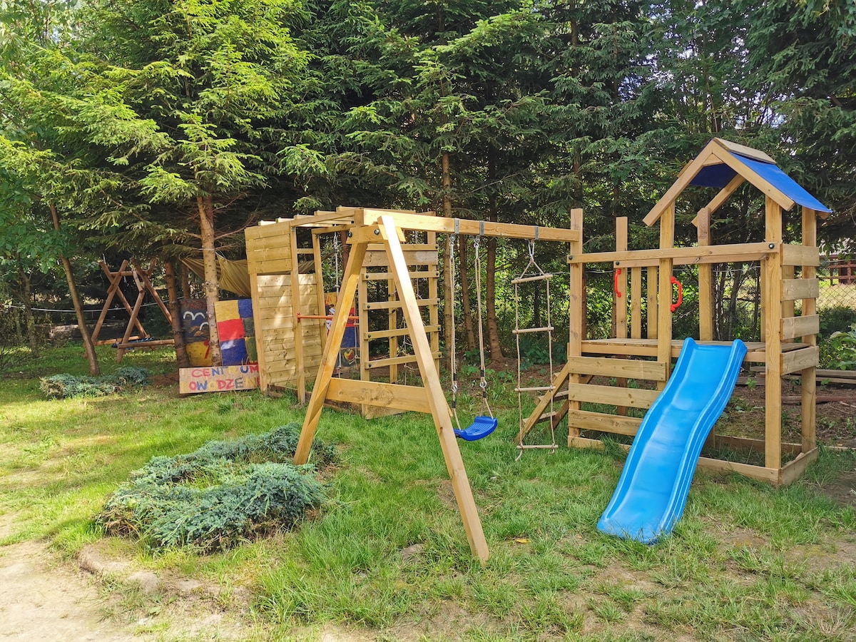 A play area is visible featuring a wooden swing set, climbing structure, and a slide. Surrounding greenery adds a natural touch, with trees providing shade. A colorful playhouse can be seen alongside a grassy patch, suitable for children's outdoor activities.