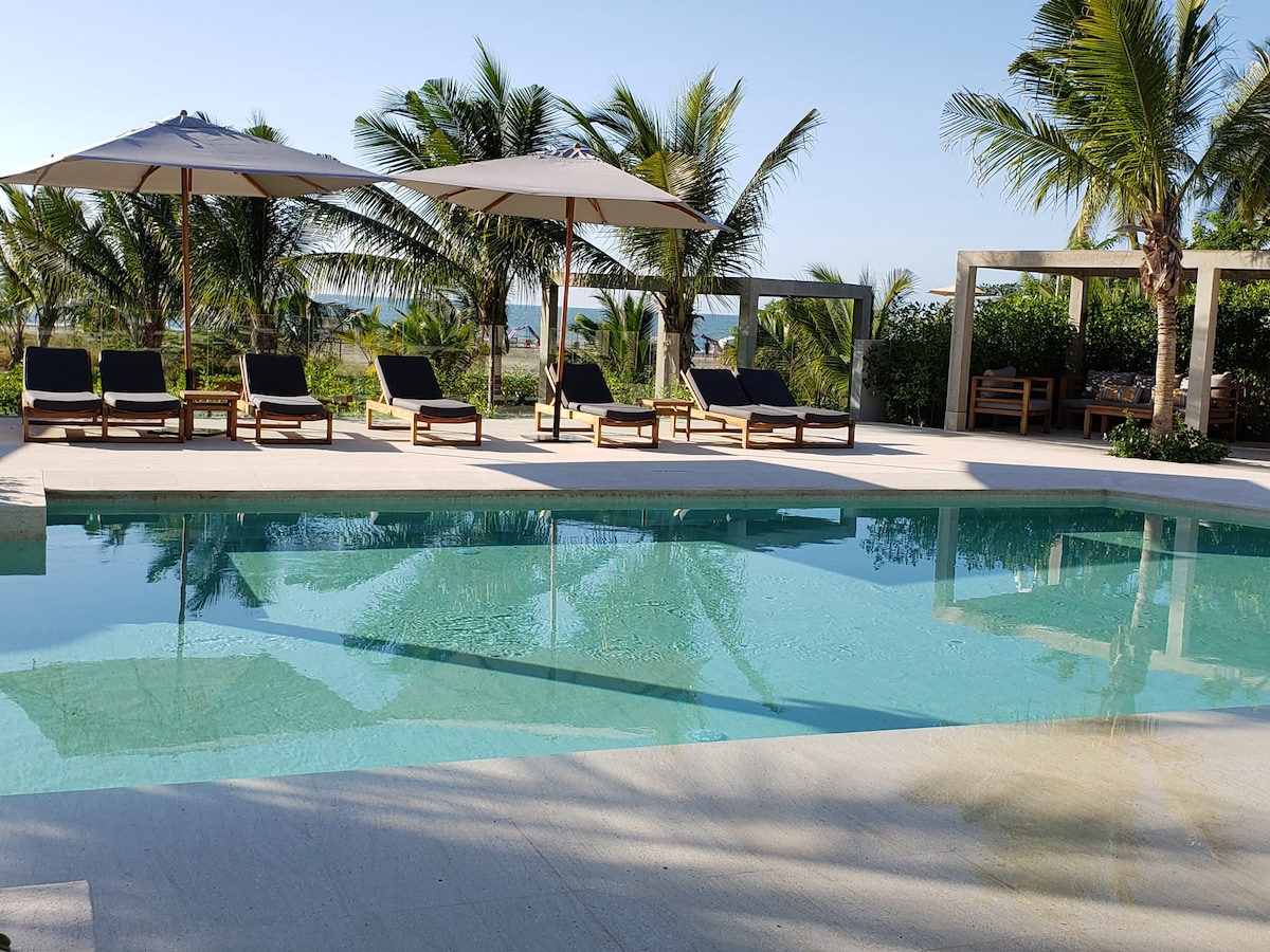 A spacious pool area is shown, featuring multiple lounge chairs arranged neatly beside the water. Large umbrellas provide shade, and palm trees are visible in the background, enhancing the outdoor relaxation space near the beach.