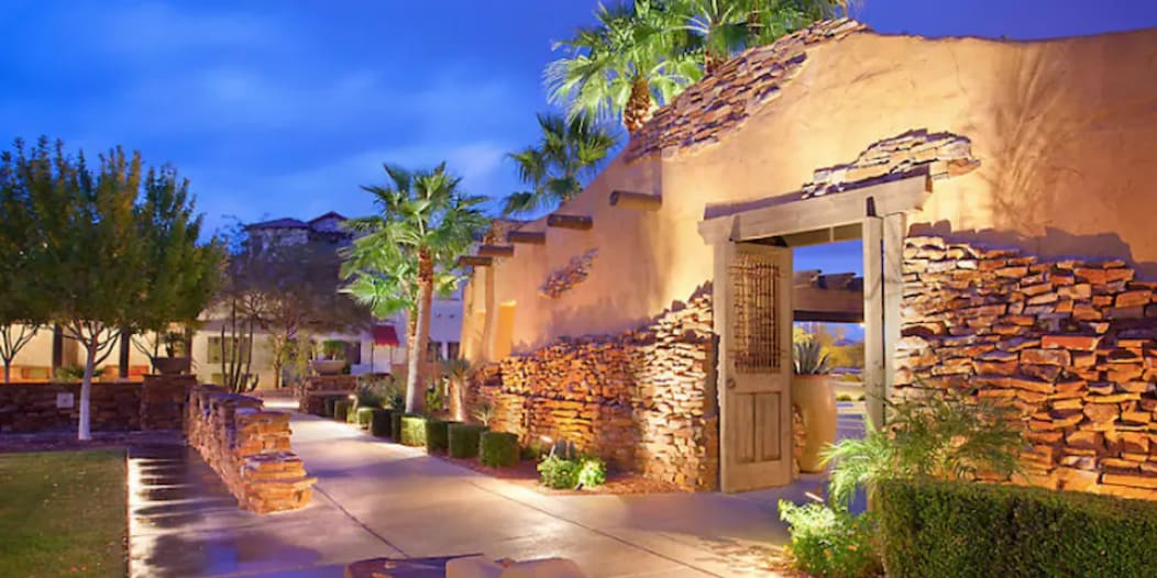 The entrance features a rustic stone wall with wooden doors, framed by well-maintained greenery. Pathways are lit softly to enhance the welcoming ambiance in the evening light, leading guests towards the resort's interior.
