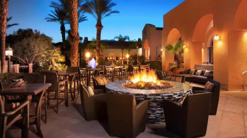 An outdoor seating area is illuminated by the warm glow of a fire pit surrounded by comfortable wicker chairs. Palm trees and soft lighting create a relaxed atmosphere, while the architectural style features arched doorways and a blend of natural elements.