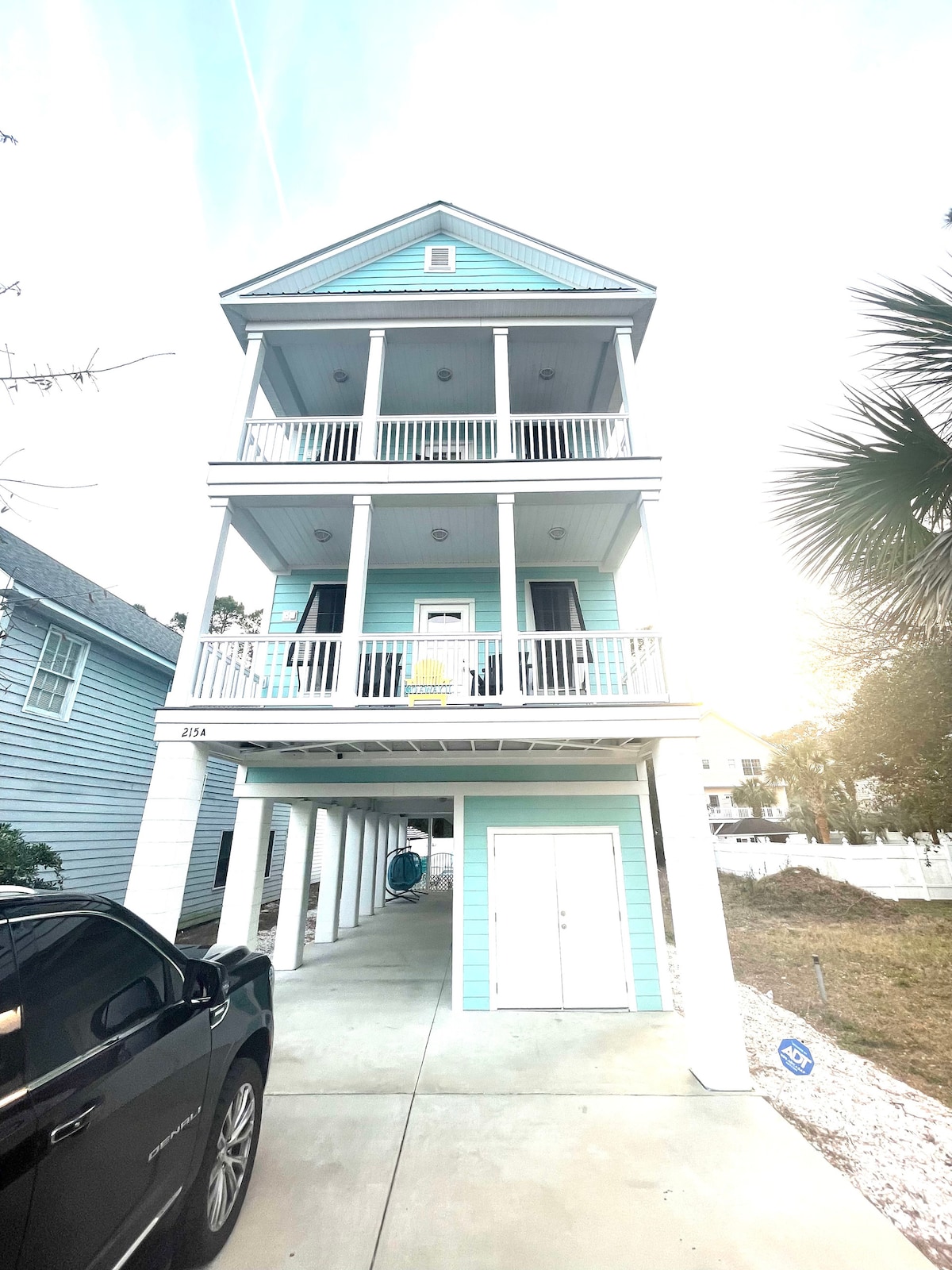 A three-story beach house is showcased, painted in light blue. The structure features multiple balconies with white railings, while a vehicle is parked in the driveway underneath. The landscaping includes palm trees and gravel, highlighting the coastal environment.