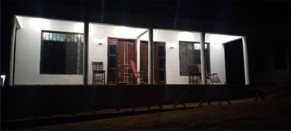 The exterior of a modern house is depicted in nighttime lighting, showcasing a wide porch with wooden chairs on either side. The entrance features double doors framed with large windows, providing a balanced and inviting aesthetic.