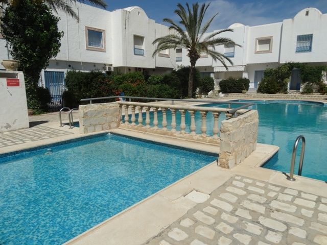 A serene pool area is featured, with clear blue water reflecting the sky. Surrounding palm trees provide a tropical vibe. A stone railing borders a smaller section of the pool, while a cobblestone walkway invites guests to enjoy the outdoor space.