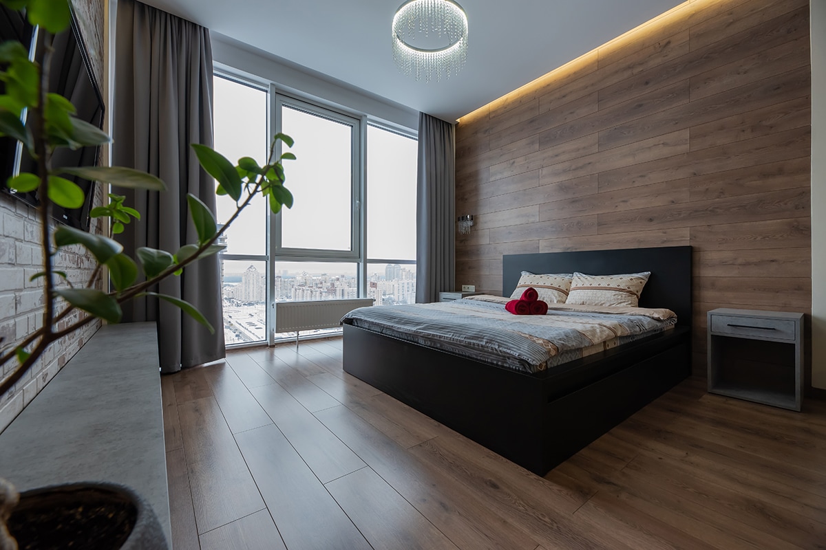 A modern bedroom features a spacious bed with neutral-toned bedding, accented by decorative pillows. Floor-to-ceiling windows allow natural light to fill the room, highlighting the wood-paneled wall. A small nightstand with a plant adds a touch of greenery to the serene atmosphere.