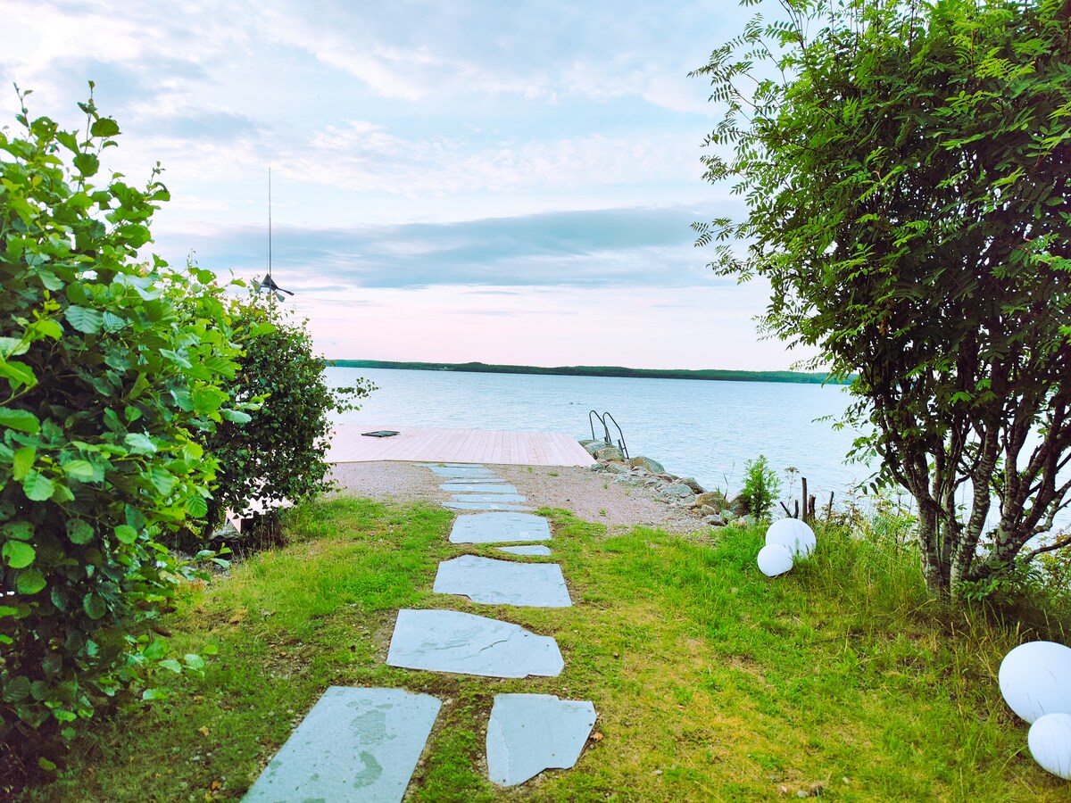 A pathway of stepping stones leads to a wooden dock by the water. Lush greenery surrounds the area, with bushes on either side and soft grass underfoot. The calm surface of the water reflects the sky, inviting exploration.