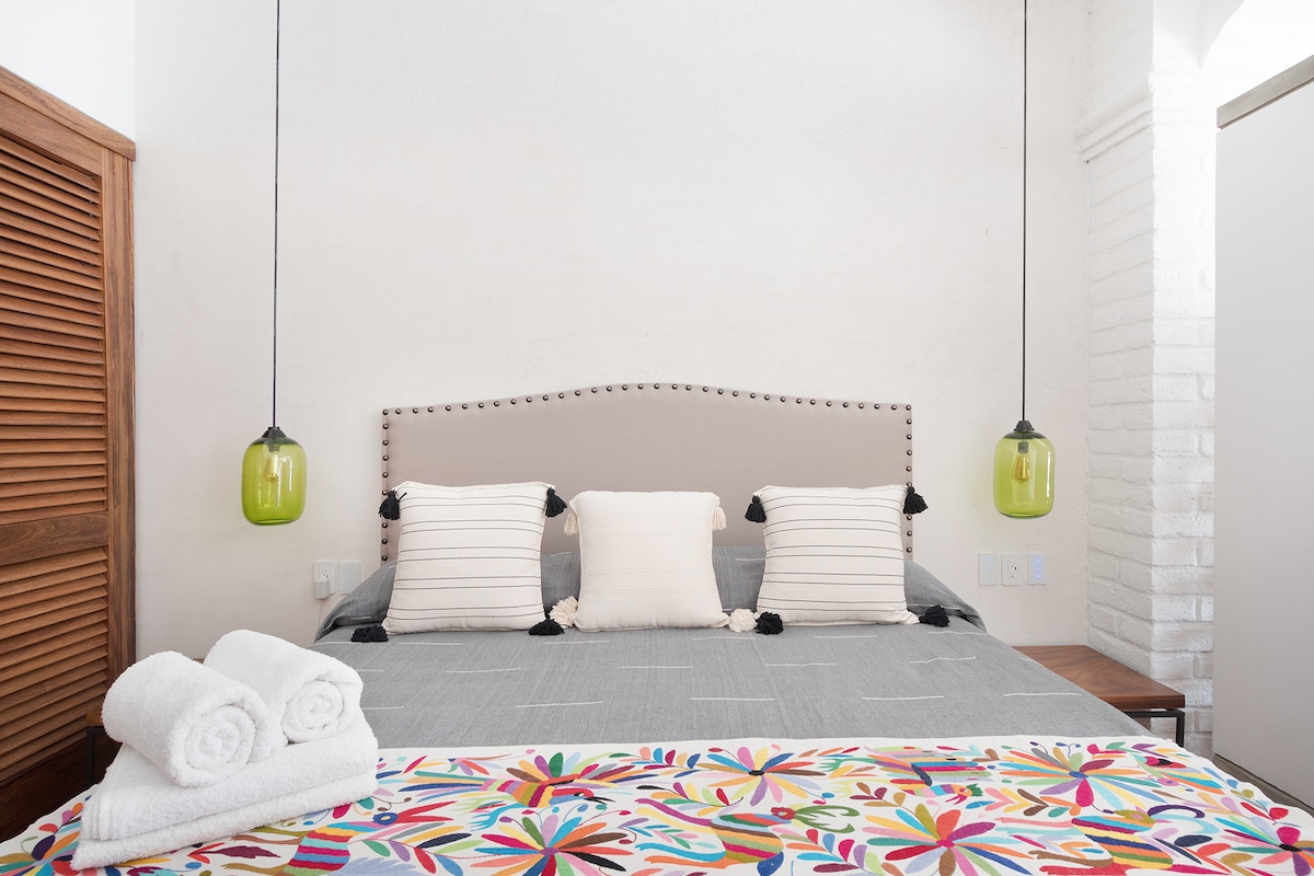 A well-appointed bedroom features a spacious bed adorned with a colorful, floral bedspread. Neutral-colored decorative pillows are placed at the head of the bed. Two pendant lights hang above, adding a modern touch, while fresh towels are neatly arranged at the foot.