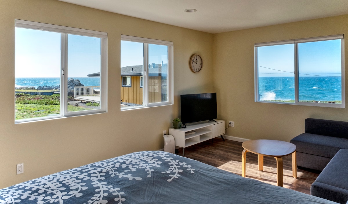 A bright and airy living space is shown, featuring multiple windows that offer views of the Pacific Ocean. A comfortable sofa and a small coffee table complement the room, while a television is positioned on a low media unit. The walls are painted a soft neutral color.