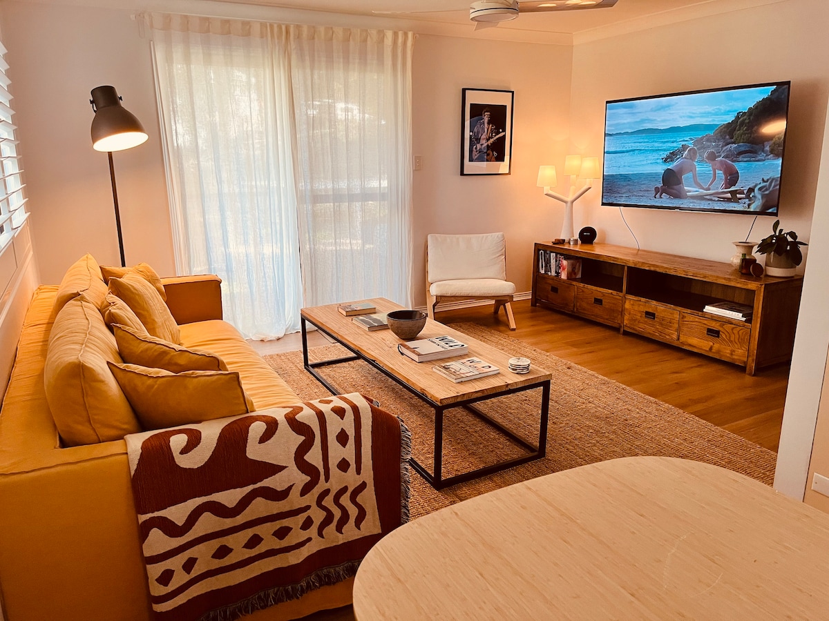 A modern living area is shown, featuring a mustard yellow sofa adorned with patterned throws. A coffee table stands in the center, surrounded by a light rug. A wall-mounted TV displays a coastal scene, and ample natural light enters through sheer curtains.