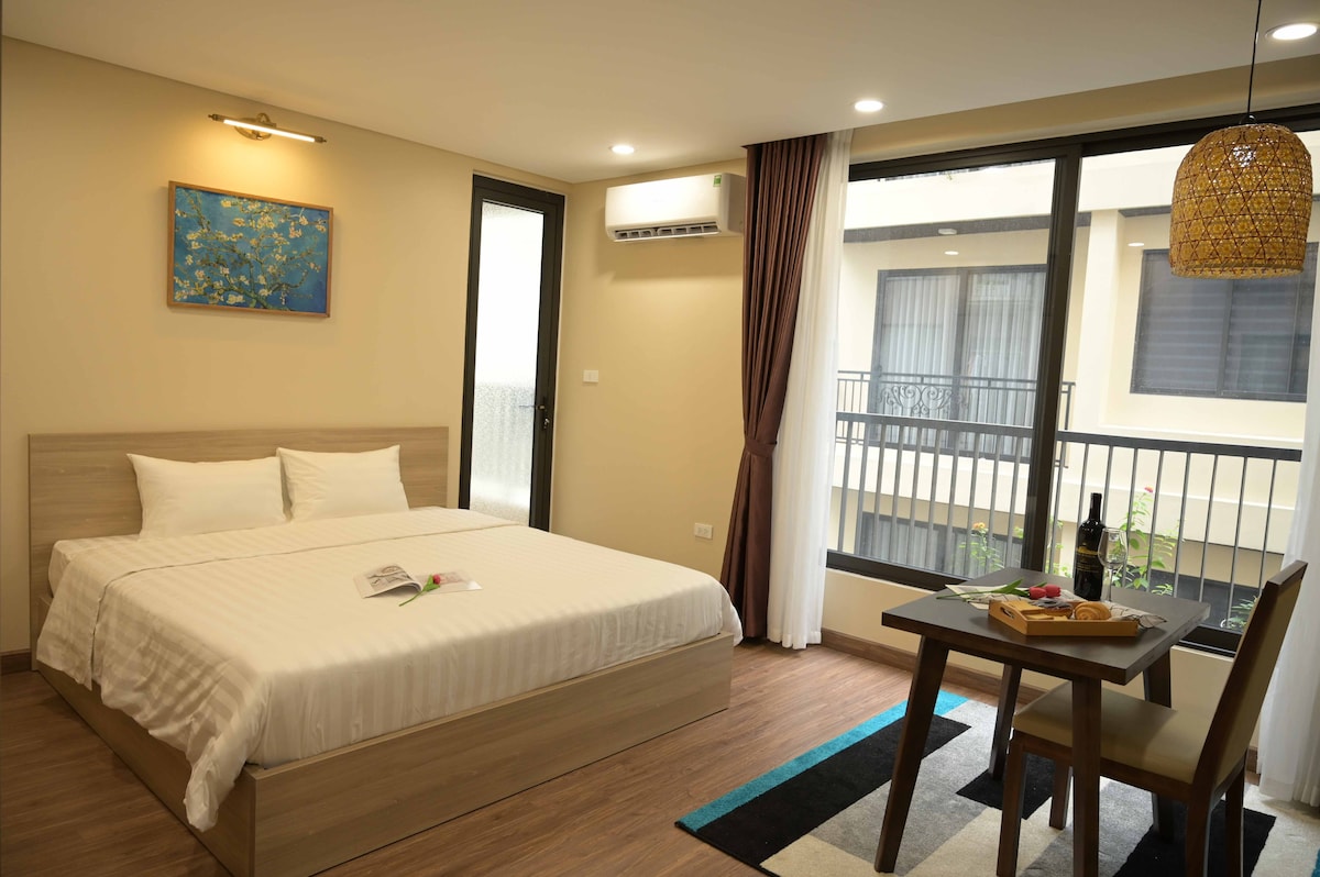 A spacious sleeping area features a large bed with crisp linens and decorative pillows. A small dining table is set nearby, with natural light streaming through large windows adorned with sheer curtains. Light wood tones and a decorative wall piece add a modern touch.