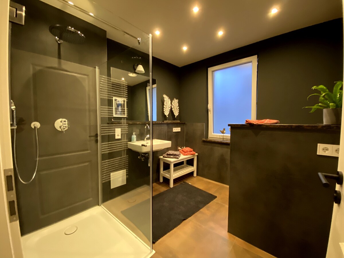 A modern bathroom is displayed, featuring a walk-in glass shower with a rain showerhead. A wall-mounted sink is visible beside a wooden shelf with neatly arranged towels. Soft lighting enhances the dark walls, while a small window allows natural light to enter.