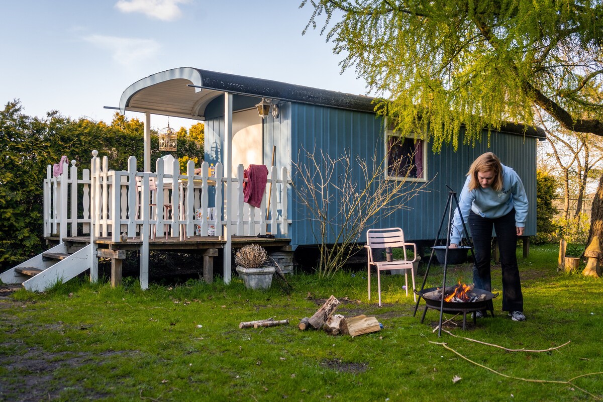 A charming pipowagen is surrounded by a grassy area, featuring a small campfire setup with logs nearby. A wooden deck with a white picket fence provides outdoor space for seating. The structure has a curved roof and simple steps leading up to the entrance.