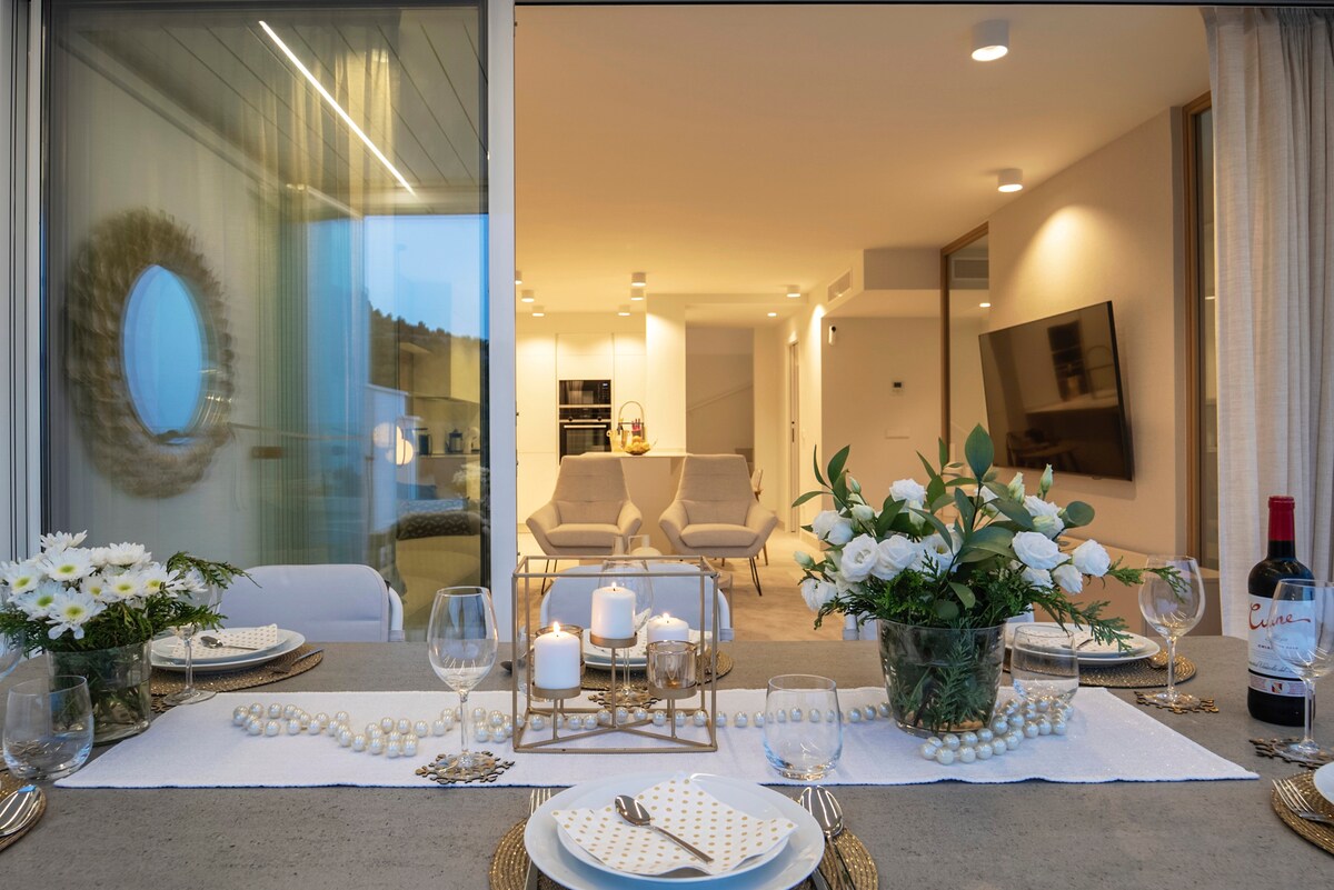 A dining table is set elegantly with plates, glasses, and a centerpiece of fresh flowers. The scene overlooks a spacious living area, featuring light-colored furniture and a large wall-mounted TV. Natural light filters through large windows, enhancing the open atmosphere.