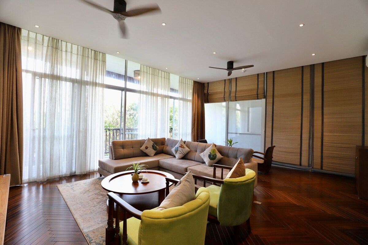 A spacious living area features large glass windows allowing natural light to fill the room. A light-colored sectional sofa complements a round wooden table, surrounded by two inviting chairs. Soft neutral curtains softly frame the windows, enhancing the open atmosphere of the space.