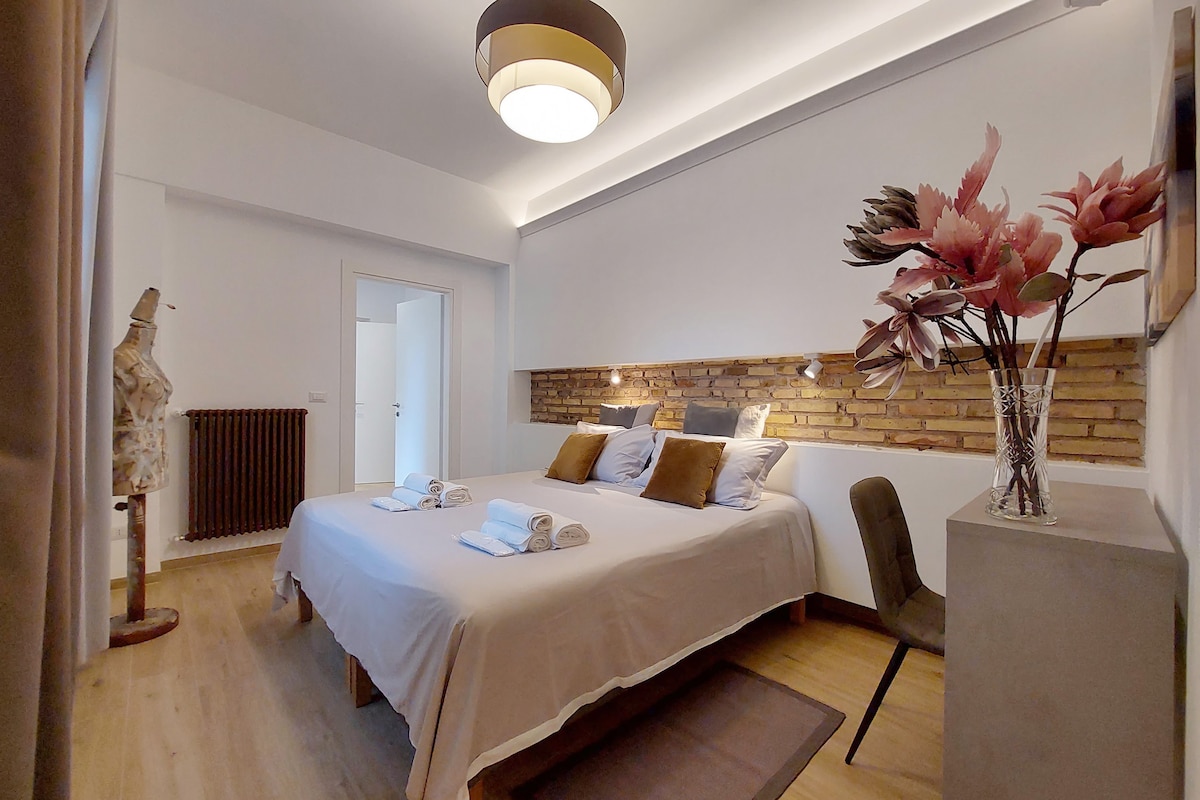 A well-appointed bedroom features a spacious bed outfitted with neutral linens and decorative cushions. Two stylish bedside tables are positioned on either side, complemented by a modern lamp. A floral arrangement adds a touch of color, while a wall with exposed brick enhances the room's character.