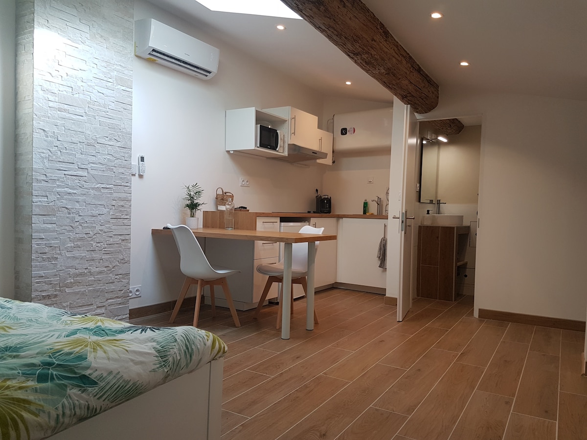 An airy studio features a cozy sleeping area with a patterned blanket on the bed. A table with two chairs occupies one corner, and a compact kitchen area includes a microwave and essential amenities. Natural light filters in through the skylight, enhancing the warm tones of the wooden flooring.