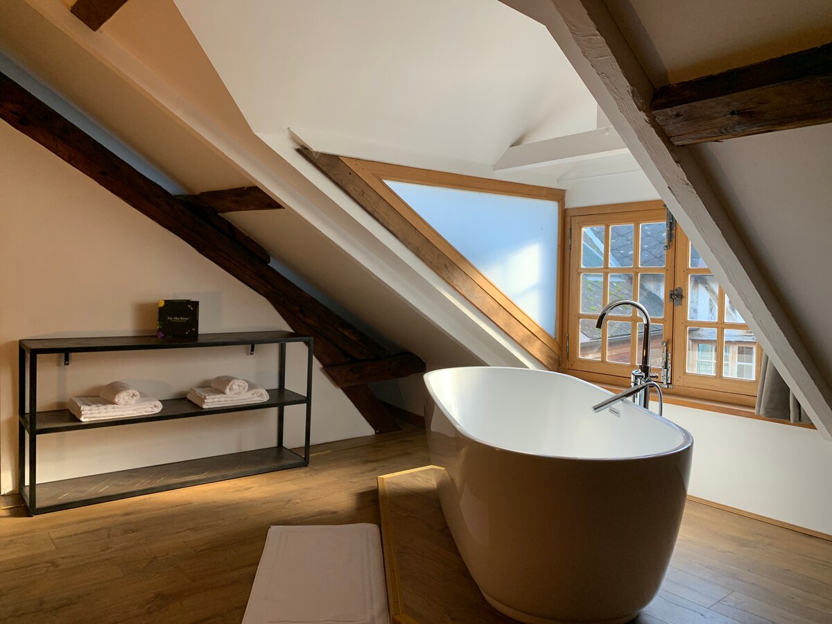 A bright bathroom features a modern freestanding tub positioned under slanted beams. Natural light fills the space through a window with wooden frames, and a shelf with neatly stacked towels is visible nearby, accentuating the room's clean and contemporary design.