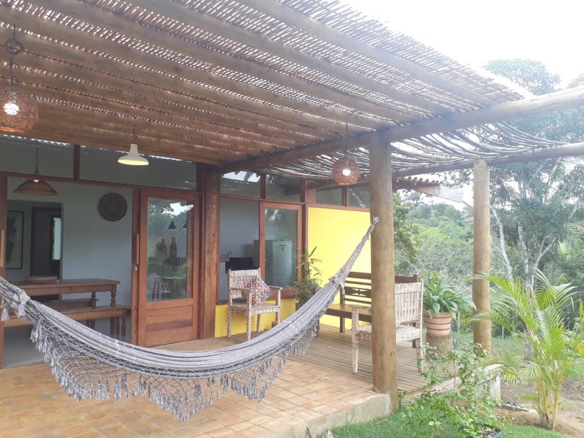 A shaded outdoor area features a woven hammock draped between wooden posts. The tan tiled floor is complemented by a wooden dining table and chairs, surrounded by lush greenery. Large glass doors lead into the interior space, allowing natural light to fill the room.