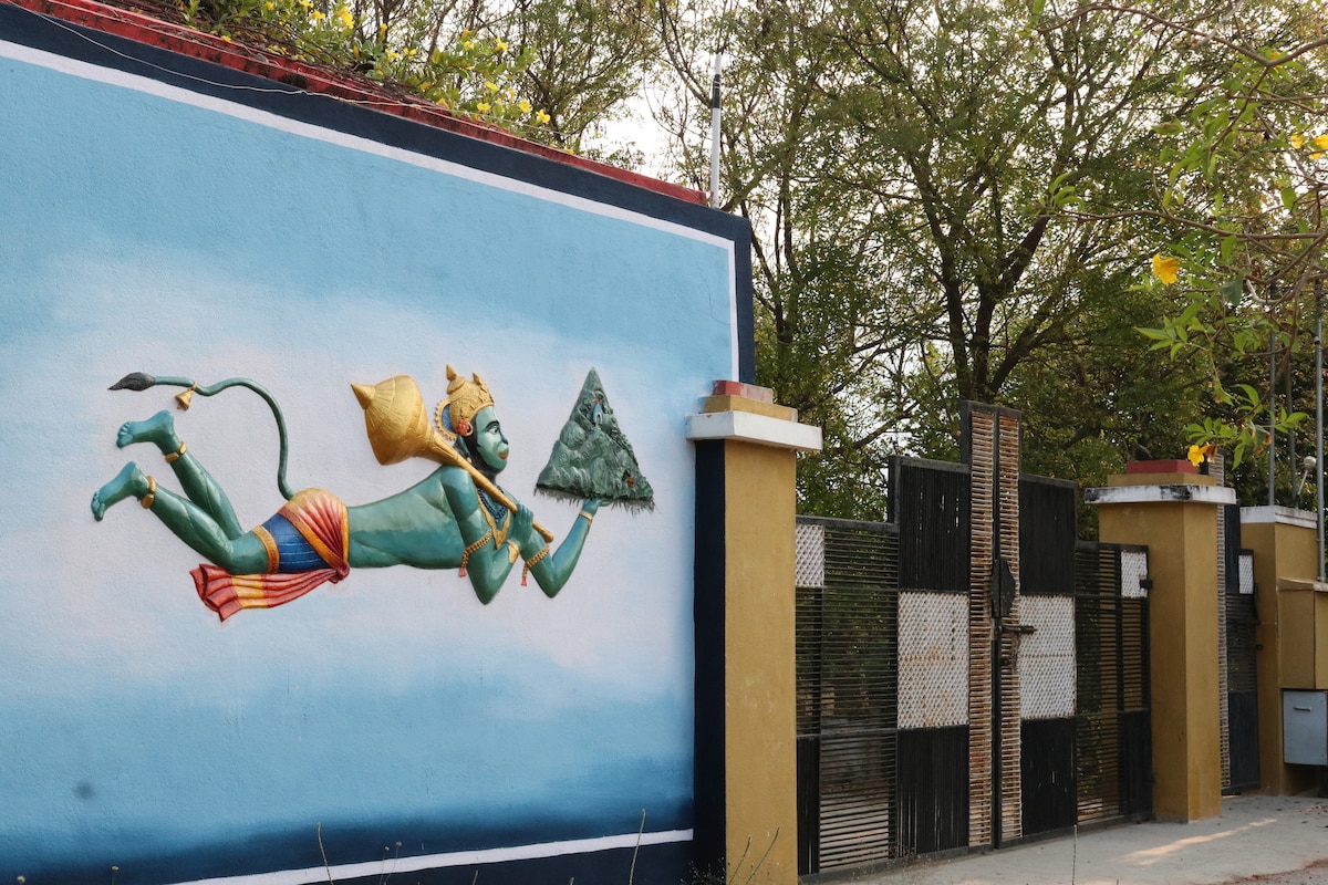 A large mural depicting a mythological figure is featured on the exterior wall. The figure is shown in mid-flight, carrying a vibrant green mountain. Surrounding trees provide a natural backdrop to the colorful artwork, enhancing the serene atmosphere of the entrance.