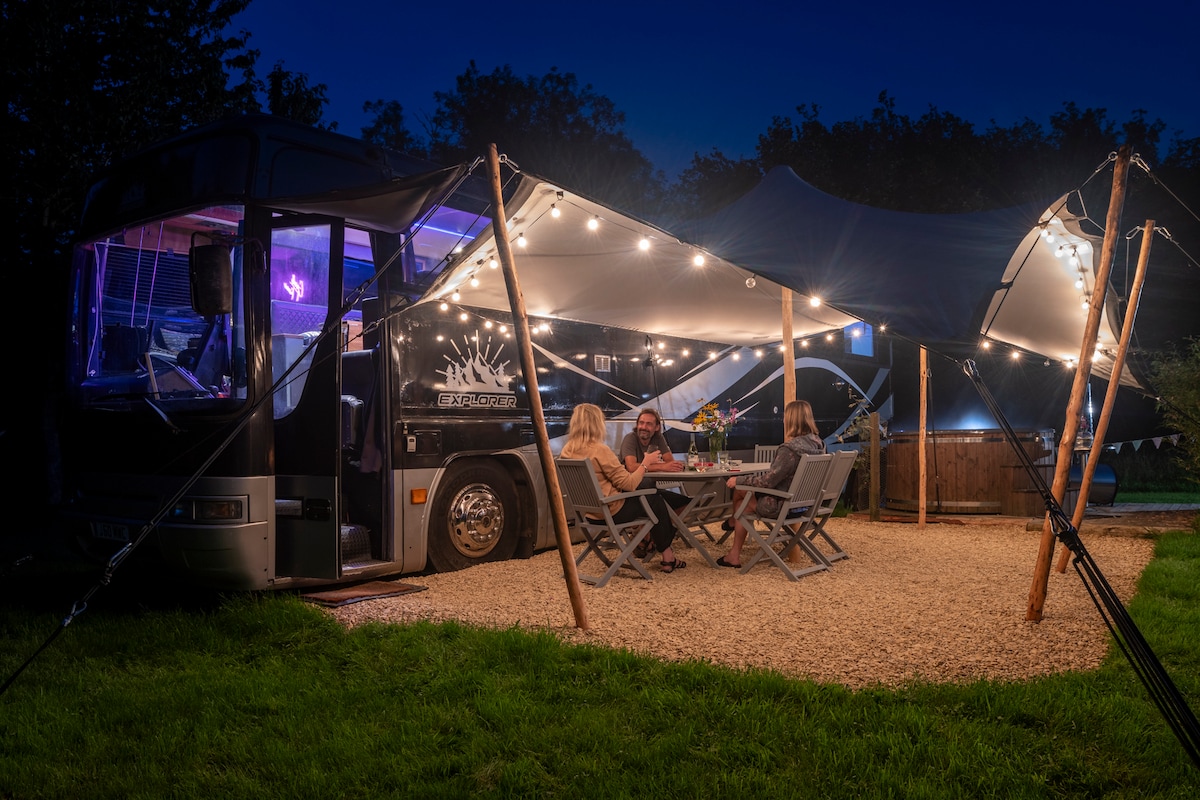 An inviting outdoor dining area is illuminated by soft string lights. A table set for four is surrounded by folding chairs on a gravel surface, with the bus and hot tub visible in the background, creating a cozy atmosphere for evening gatherings.