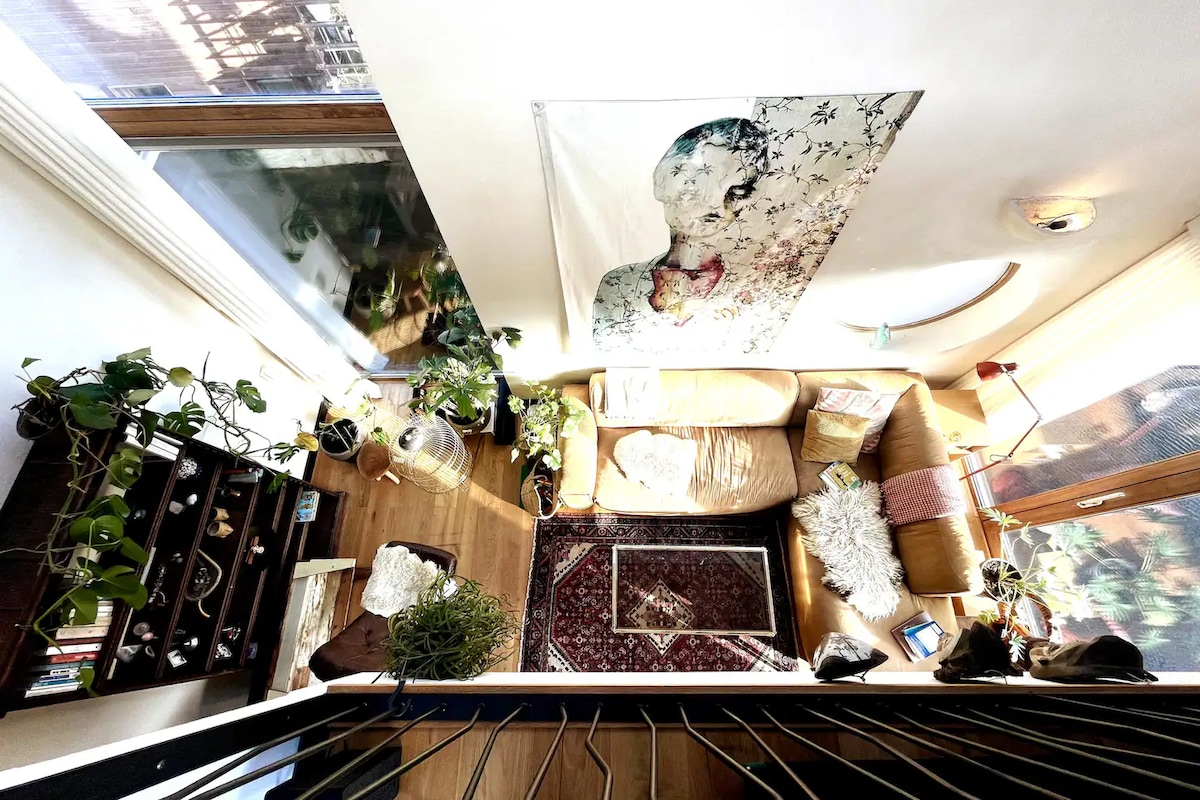An inviting living area is displayed from an overhead perspective, featuring a comfortable couch complemented by a patterned area rug. Green plants are positioned throughout the space, and a large artwork adorns the wall, enhancing the calm ambiance. Natural light enters through large windows.