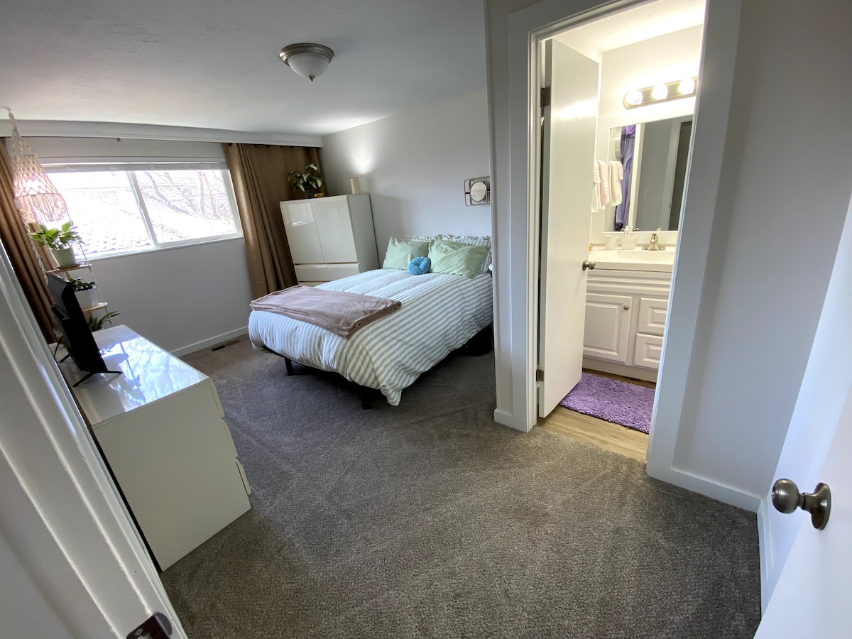 A well-lit private bedroom features a full size bed with light bedding. A white dresser stands across from the bed, and natural light enters through the window covered by sheer curtains. An attached bathroom is visible, showcasing a clean and organized layout.