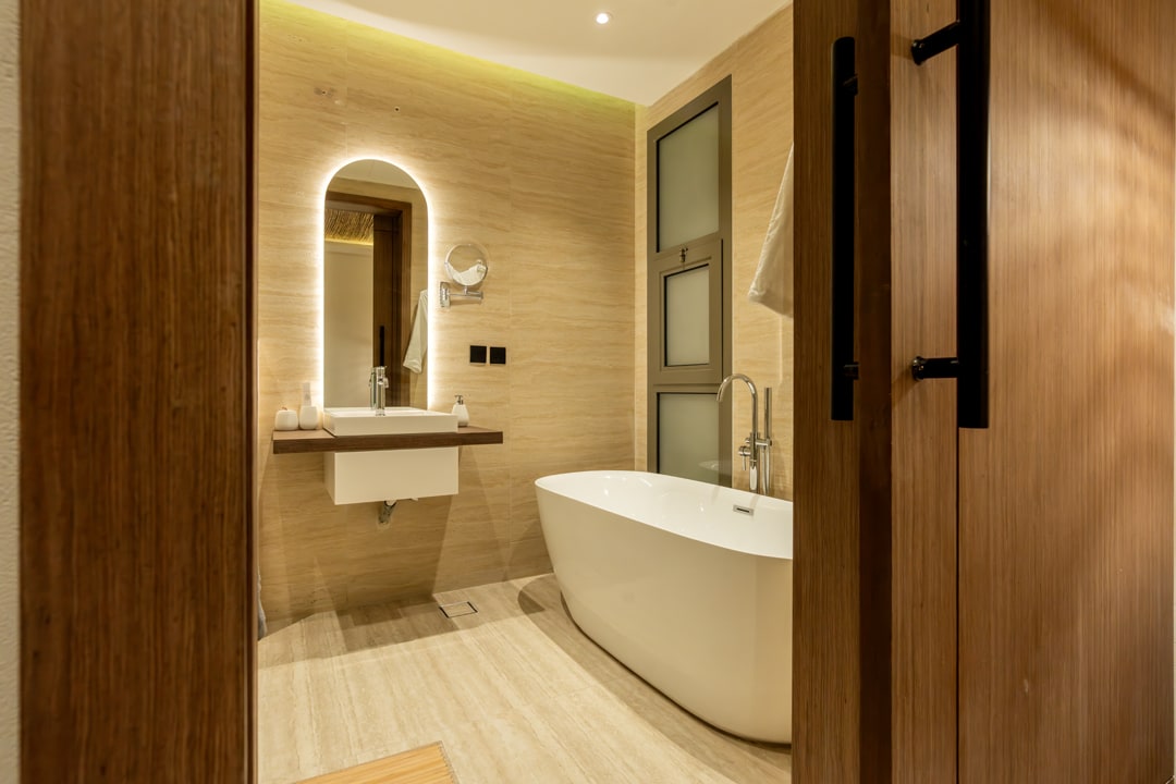 A modern bathroom is featured, with a freestanding bathtub positioned against a natural stone wall. A wooden vanity holds a round mirror and a sink, complemented by soft lighting. Simple, elegant fixtures enhance the room's clean design, creating a serene ambiance.