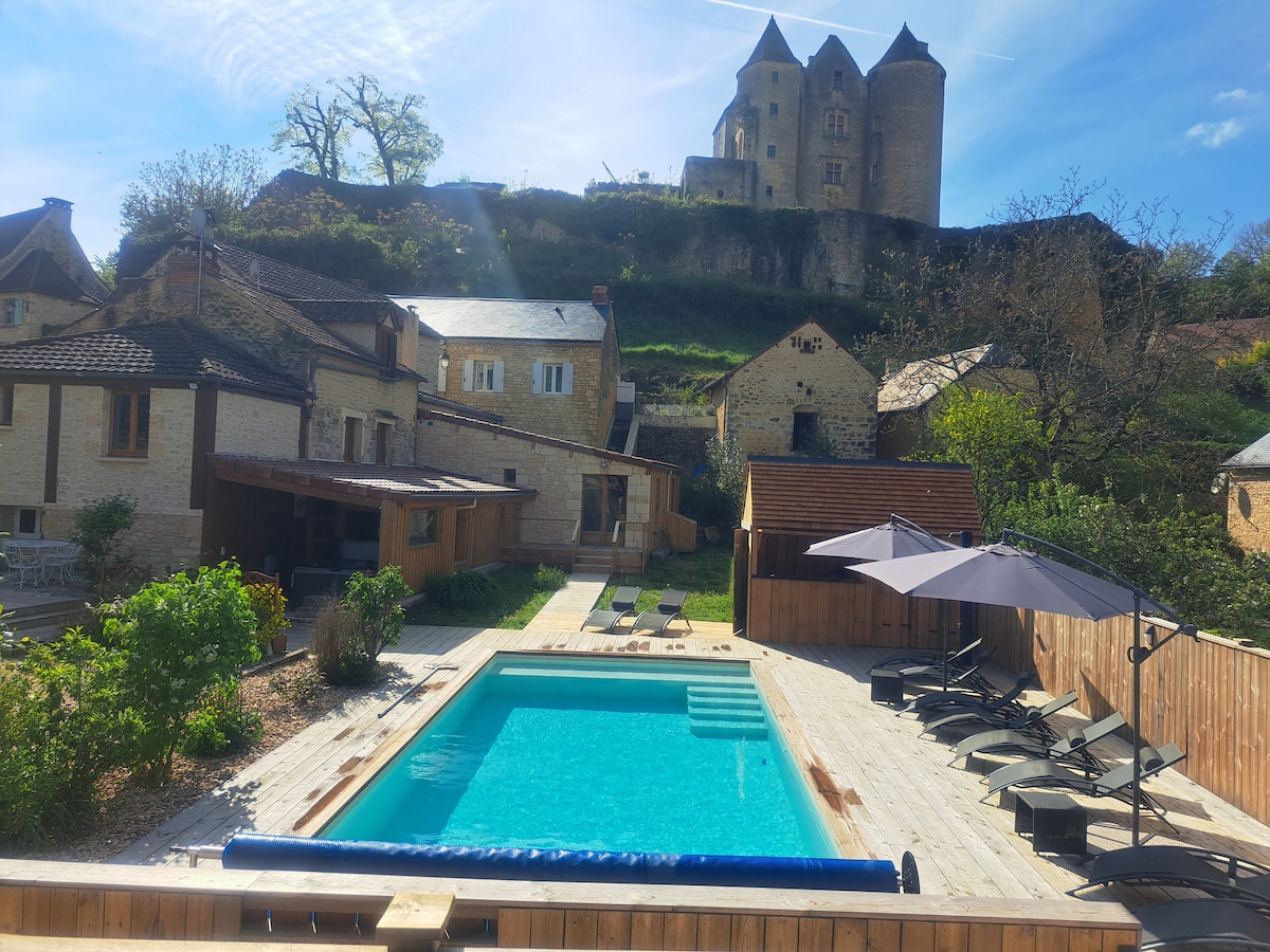 A secure outdoor pool is surrounded by a wooden deck, featuring lounge chairs and umbrellas. In the background, a historic castle looms above lush greenery, adding to the tranquil setting. The area is well-lit with natural light, suggesting a peaceful atmosphere.