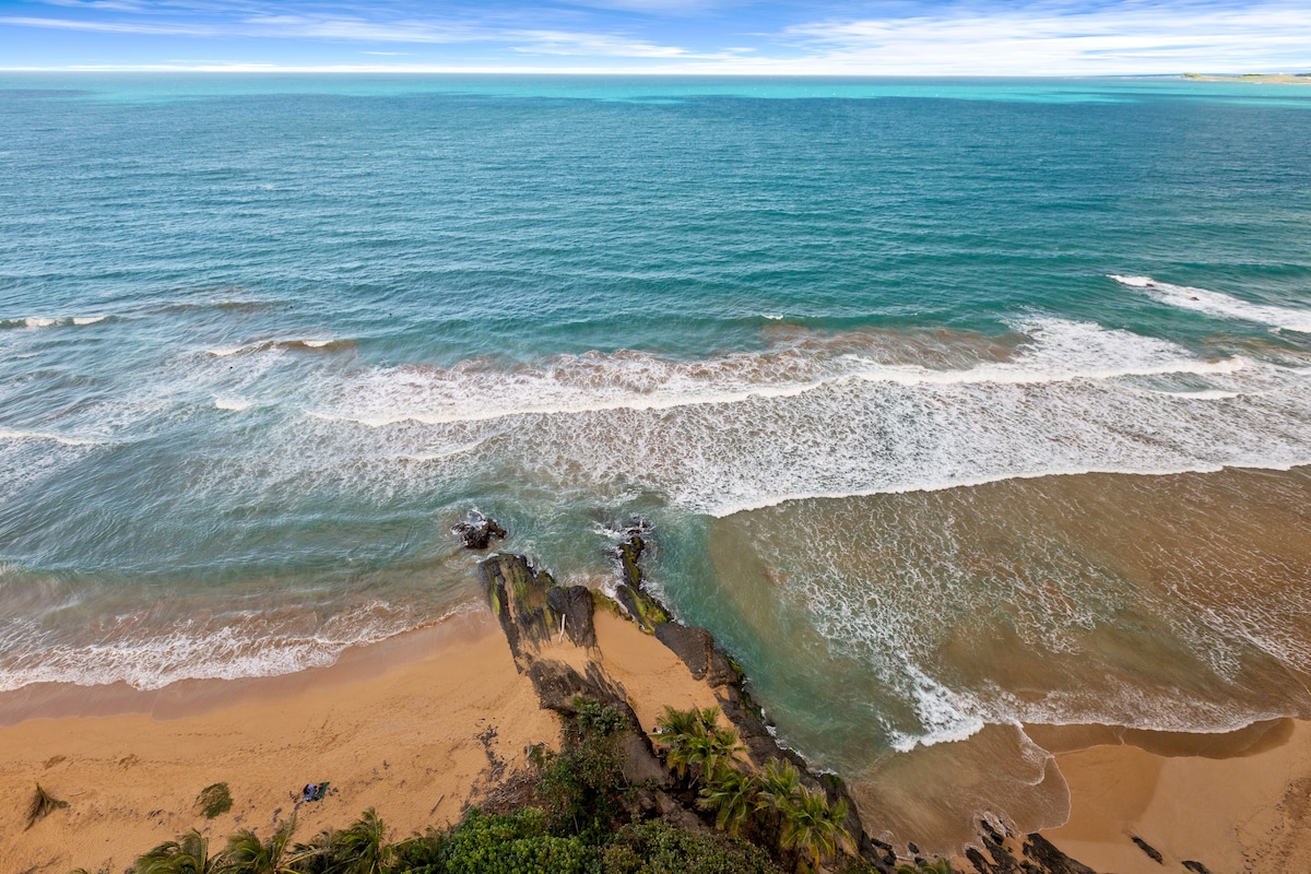 An expansive view captures the ocean meeting a sandy beach, showcasing gentle waves lapping at the shore. Rocks jut out from the coastline, partially covered in greenery, under a clear blue sky. The horizon fades into varying shades of blue, highlighting the vast sea.
