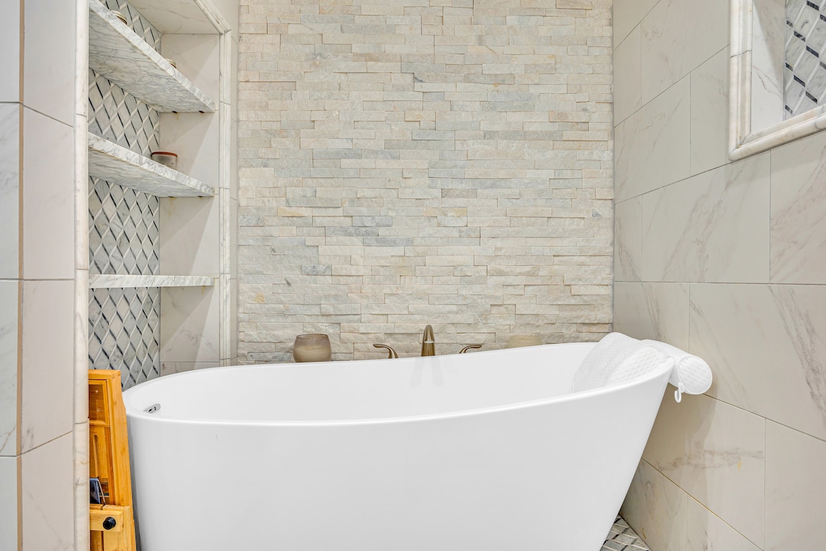 A freestanding white bathtub is positioned against a textured stone wall, offering a serene bathing experience. Shelving units are built into the wall, providing space for toiletries. Elegant fixtures are featured near the bathing area, enhancing a modern aesthetic.