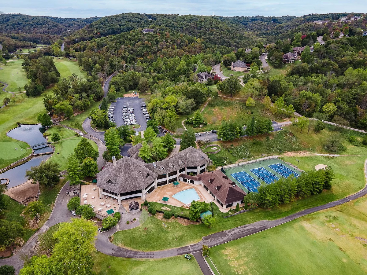 An aerial view captures the expansive grounds of the Roark Lodge, surrounded by lush greenery and rolling hills. The lodge features a swimming pool and several tennis courts. Nearby, a paved road winds through the community, leading to ample parking and landscaped areas.