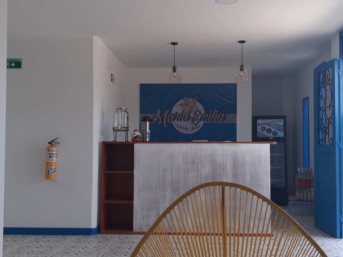 A welcoming reception area features a wooden front desk with a logo display. Two pendant lights provide illumination. The space is enhanced by a beverage dispenser and a visible storage unit. The floor showcases decorative tiles, contributing to a modern aesthetic.
