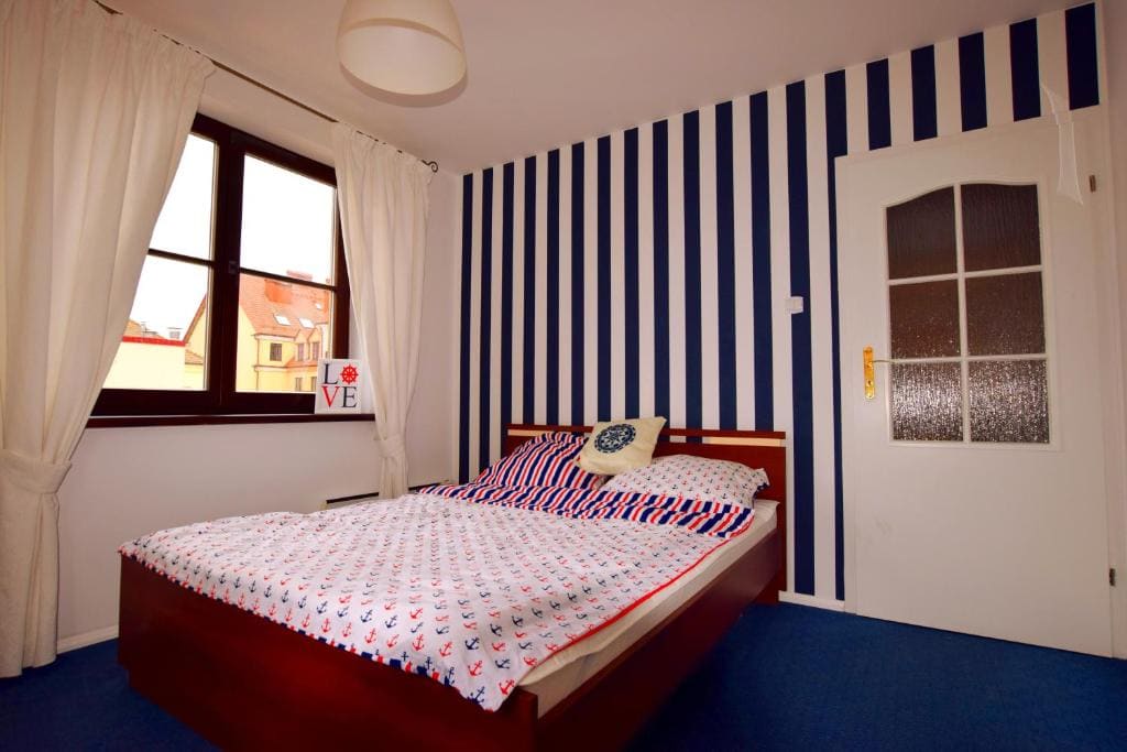 A cozy bedroom is presented, featuring a spacious bed adorned with a patterned duvet cover and throw pillows. Striped blue and white walls complement the room, while a large window invites natural light. A simple door with a frosted glass panel is visible.