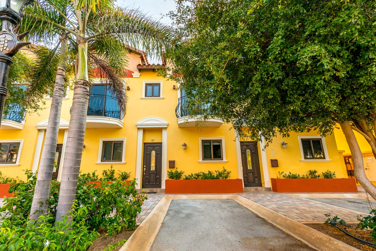 The exterior of the townhouse features vibrant yellow walls and dark wooden doors. Lush greenery and palm trees frame the entrance, while spacious walkways and balconies add to the inviting atmosphere of the property.