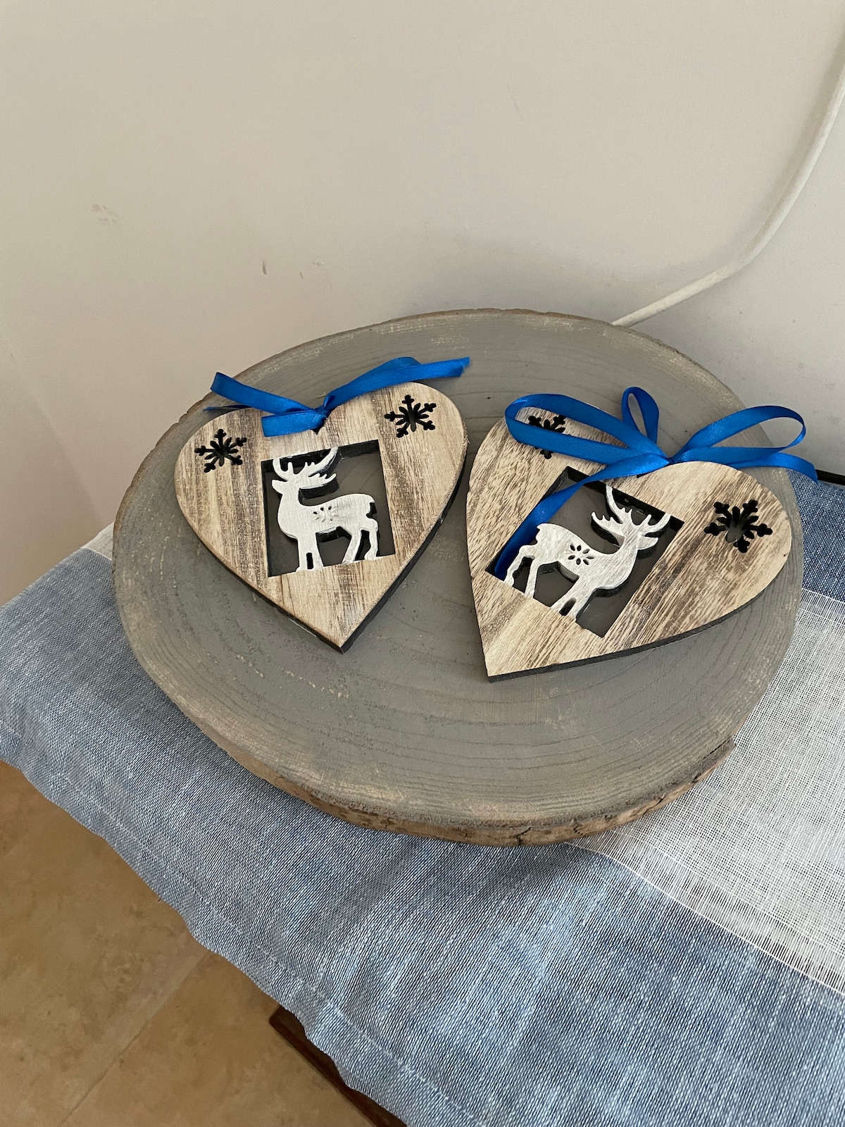 Two heart-shaped wooden ornaments are displayed on a round gray plate. Each ornament features a cut-out design of a reindeer, complemented by a blue ribbon tied at the top. The background shows a textured tablecloth with blue and white stripes.