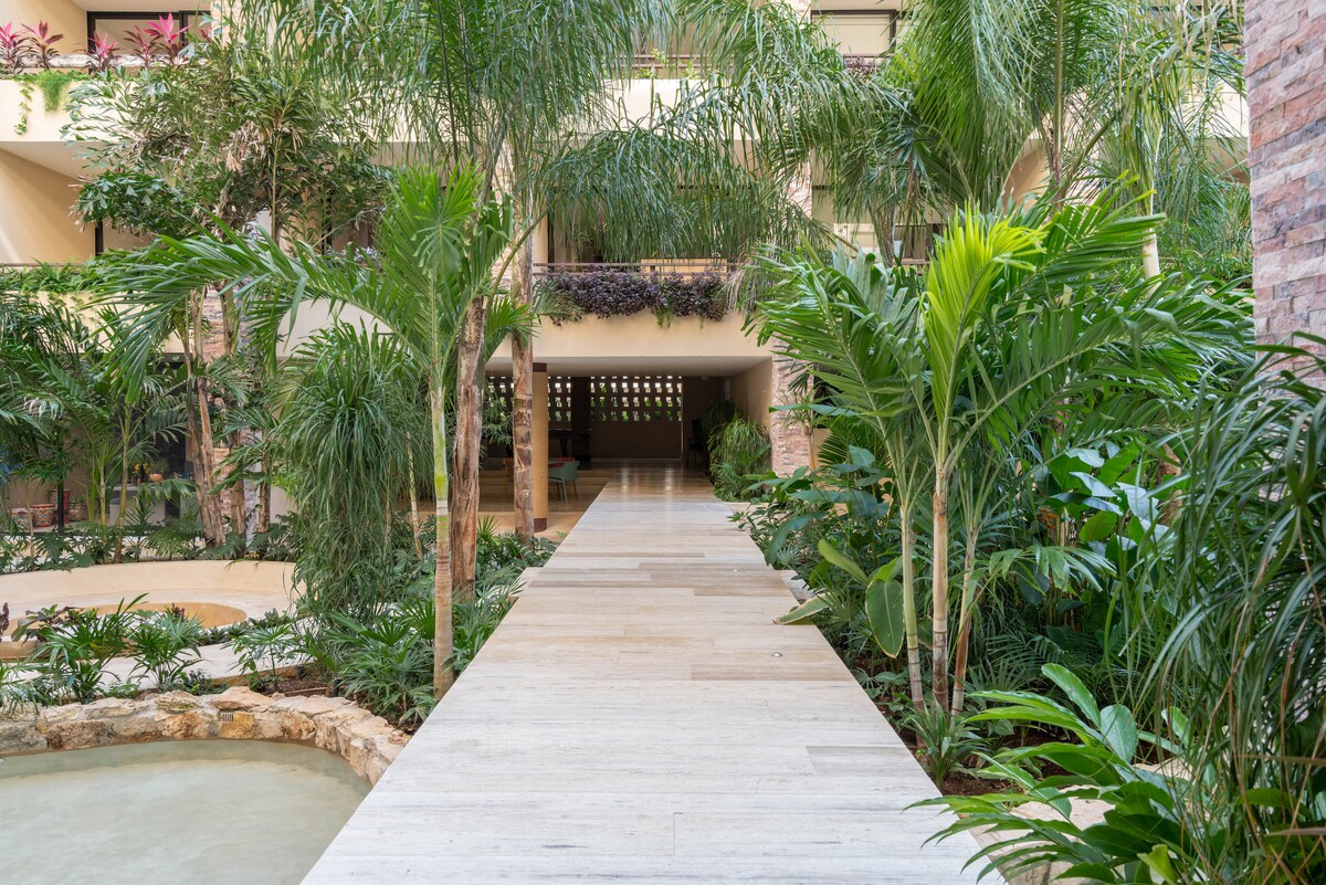 A serene pathway of light-colored stone extends through lush tropical gardens. Various palm and leafy plants surround the walkway, creating a calm atmosphere. Soft, natural light filters through the greenery, accentuating the tranquil environment leading towards the entrance.