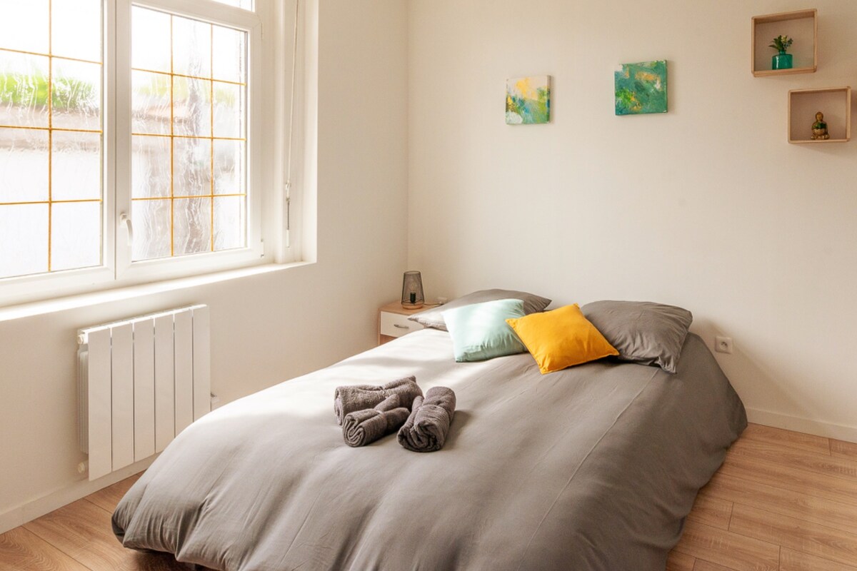 The sleeping area features a spacious bed adorned with soft pillows, one in yellow and others in hues of green. A light blanket covers the bed, and a pair of neatly rolled towels is placed on top. Natural light enters through the large windows.