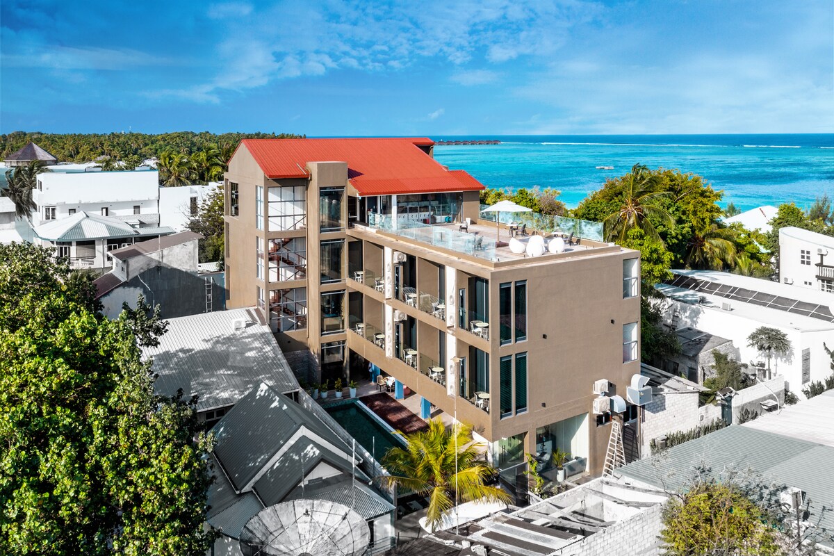 The hotel building is illustrated from an aerial perspective, featuring a distinctive red roof. Surrounding greenery and neighboring structures are visible, along with views of the ocean beyond. Balconies equipped with furniture are present, enhancing the sense of open space.