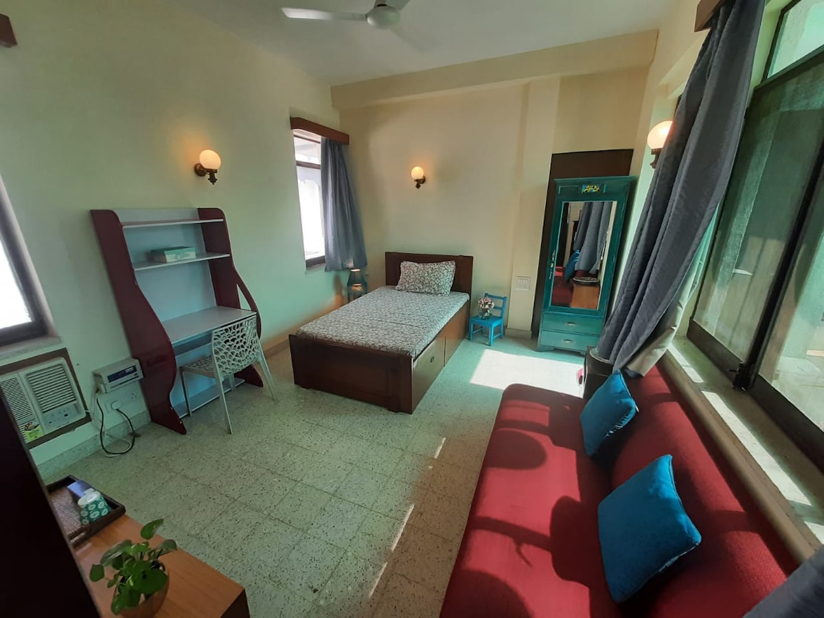 A single room features a spacious bed dressed in light bedding, flanked by two windows allowing natural light. A red sofa with cushions complements a small wooden table. A light grey study desk and a blue wardrobe are positioned against the walls, creating a functional living space.
