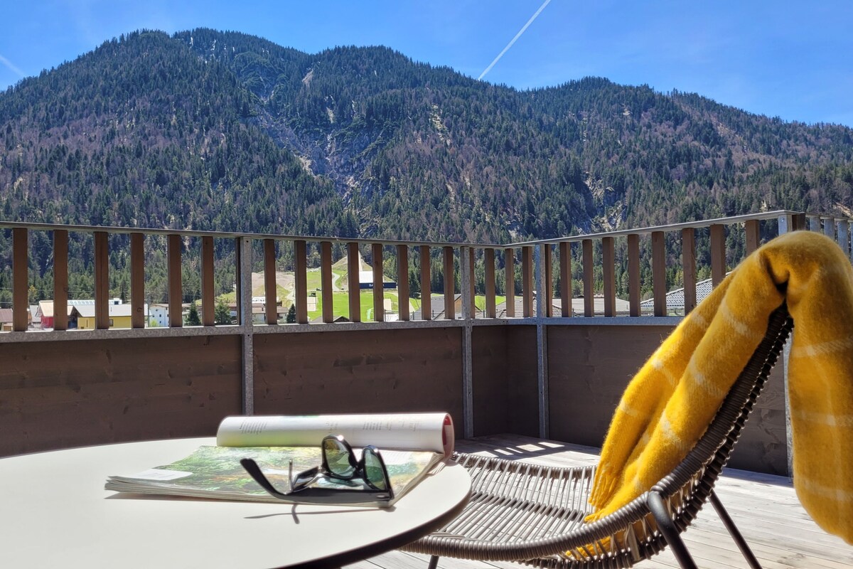 A balcony offers a scenic view of mountain peaks and green valleys. A small table displays a rolled map and sunglasses, while a cozy yellow blanket drapes over a modern chair, suggesting a comfortable spot to relax and appreciate the surrounding landscape.