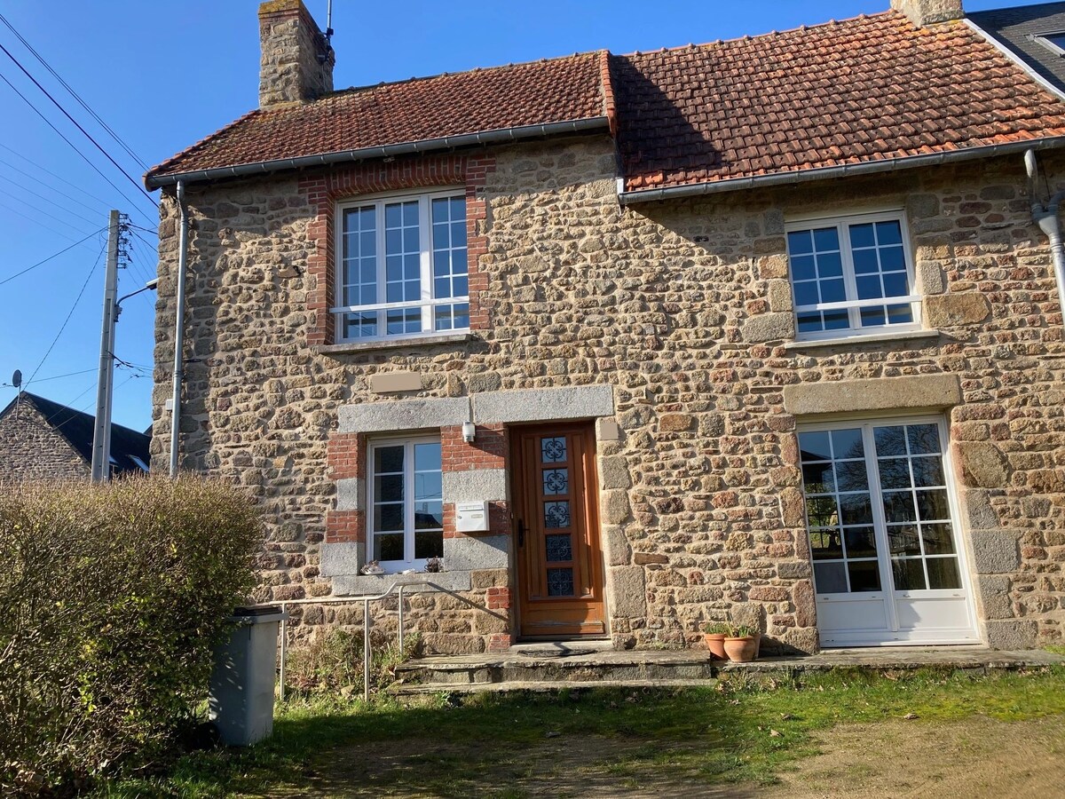 A charming stone house features a sloped roof and large windows that allow natural light to fill the interior. The wooden front door is framed by red brick, and a small set of stairs leads to the entrance. Surrounding greenery softens the façade.
