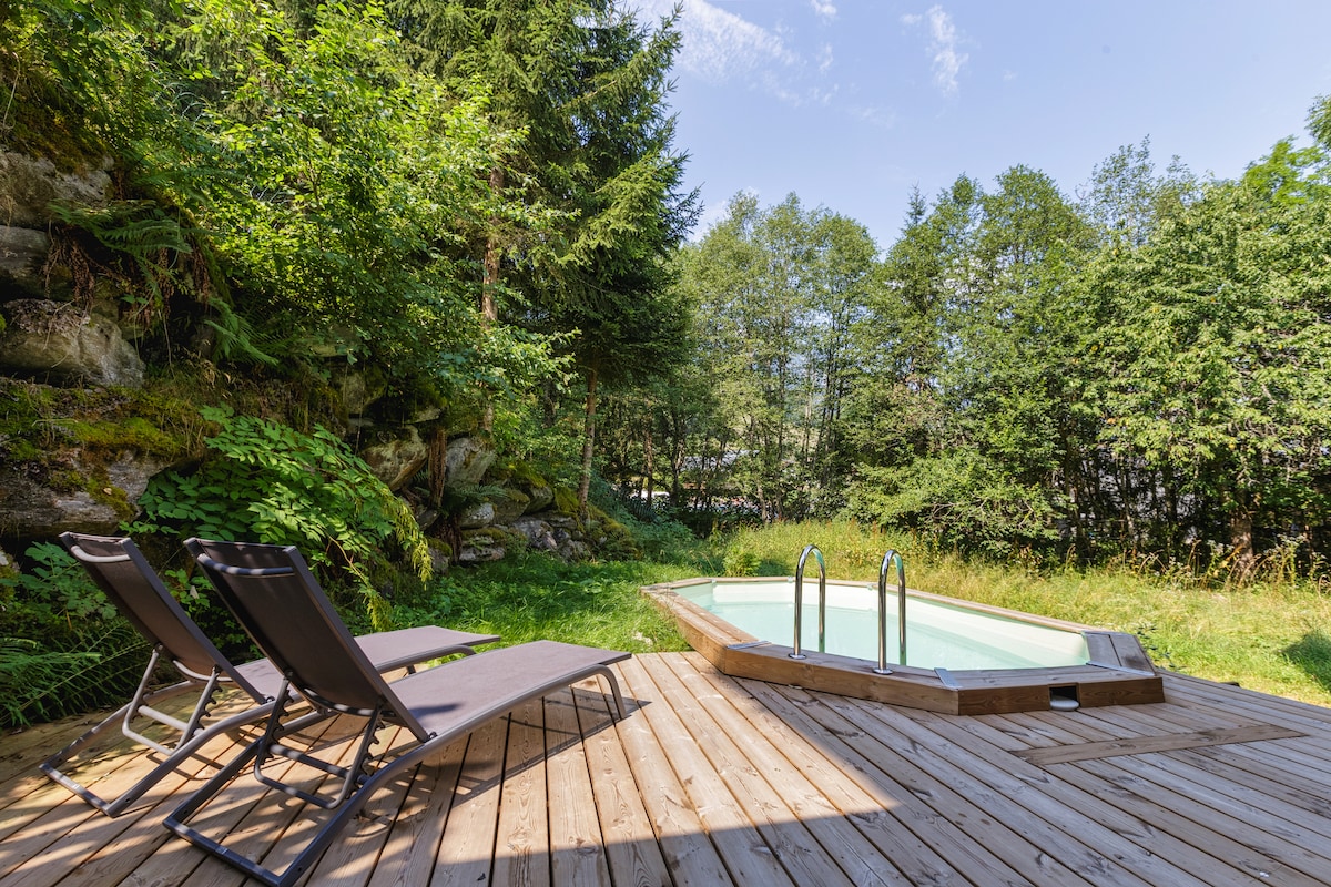 A tranquil outdoor area features a private pool surrounded by lush greenery. Two loungers sit on a wooden deck, offering a relaxing space for enjoyment. The natural ambiance is complemented by the surrounding trees, creating a serene escape.