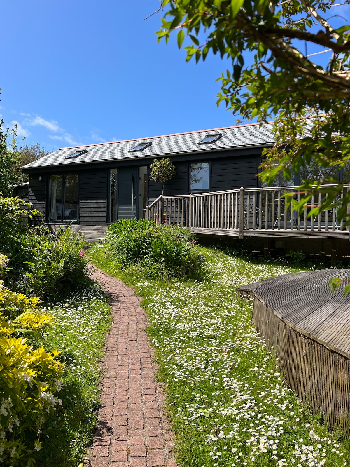 The cabin is set amidst lush greenery, featuring a winding brick pathway leading to the entrance. A wooden deck extends around the cabin, offering a spacious outdoor area. Bright flowers and foliage surround the path, enhancing the serene garden atmosphere on a clear blue sky day.